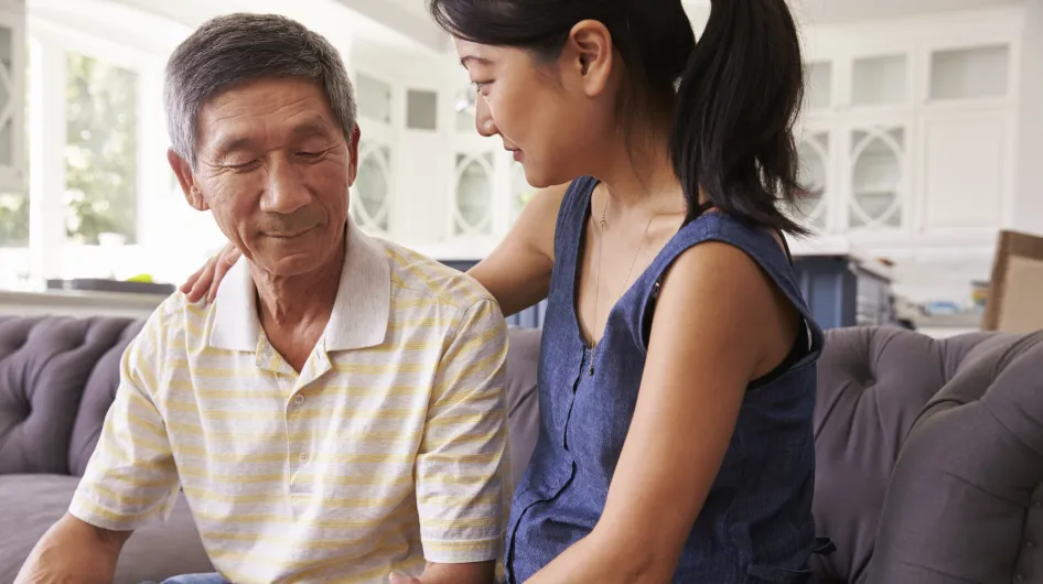 A young Asian woman sits on the couch with her elderly father having a conversation